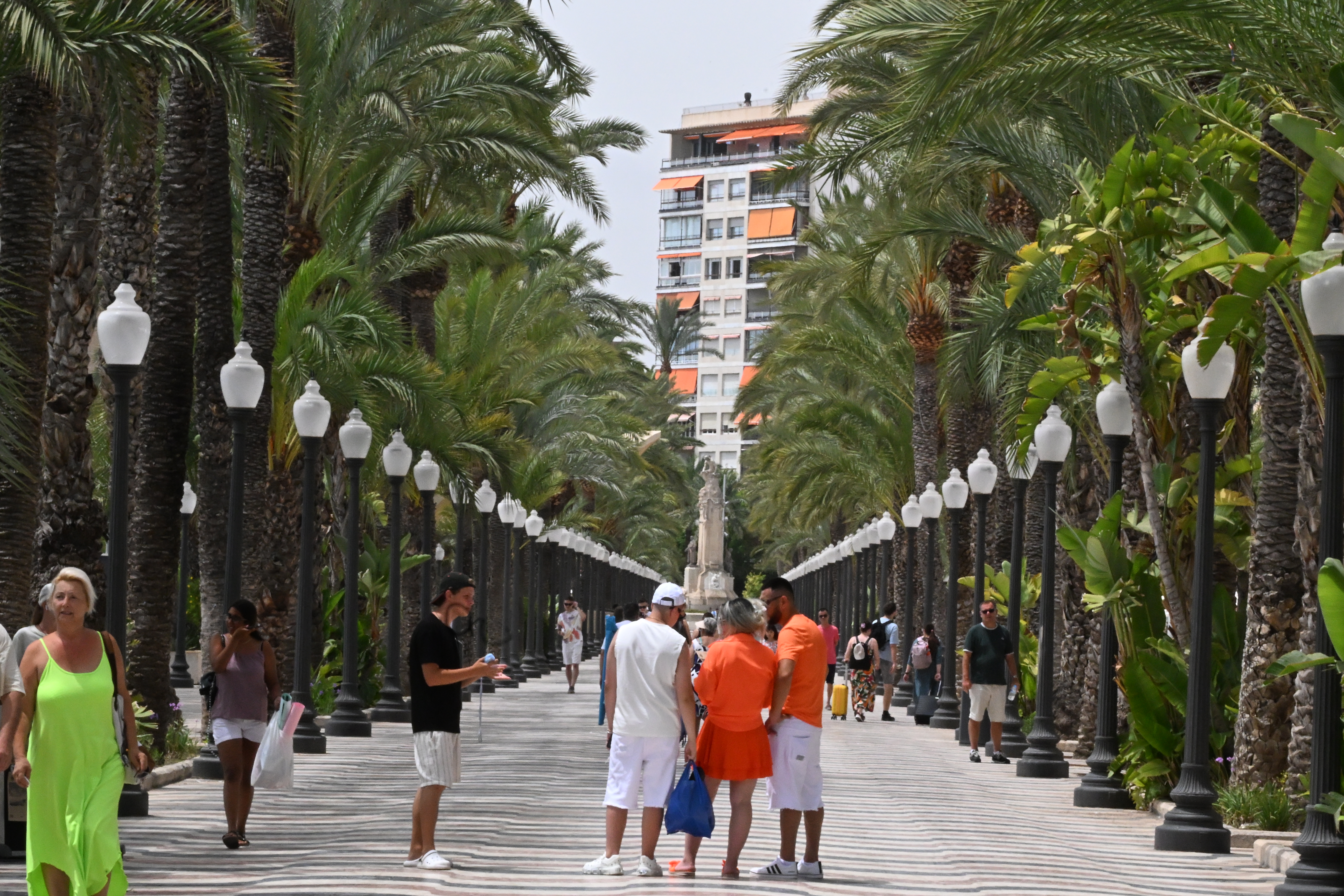 Passanten auf der palmengesäumten Explanada de España in Alicante, mit Laternenreihen und dem Monumento a Canalejas in der Ferne.