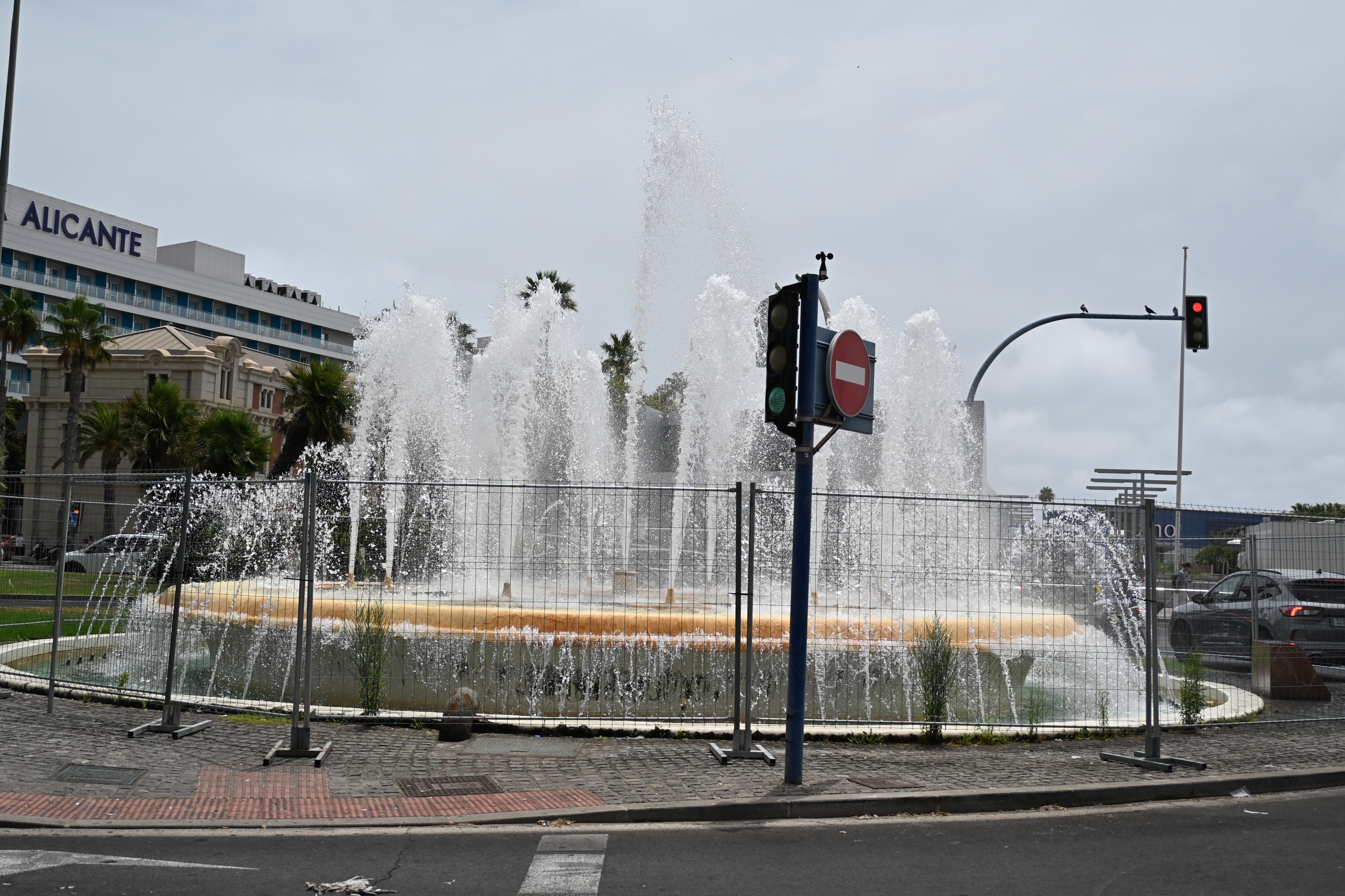 Hohe Wasserstrahlen der runden Fontäne an der Puerta del Mar in Alicante, umgeben von Absperrgittern, Palmen und Straßenverkehr.