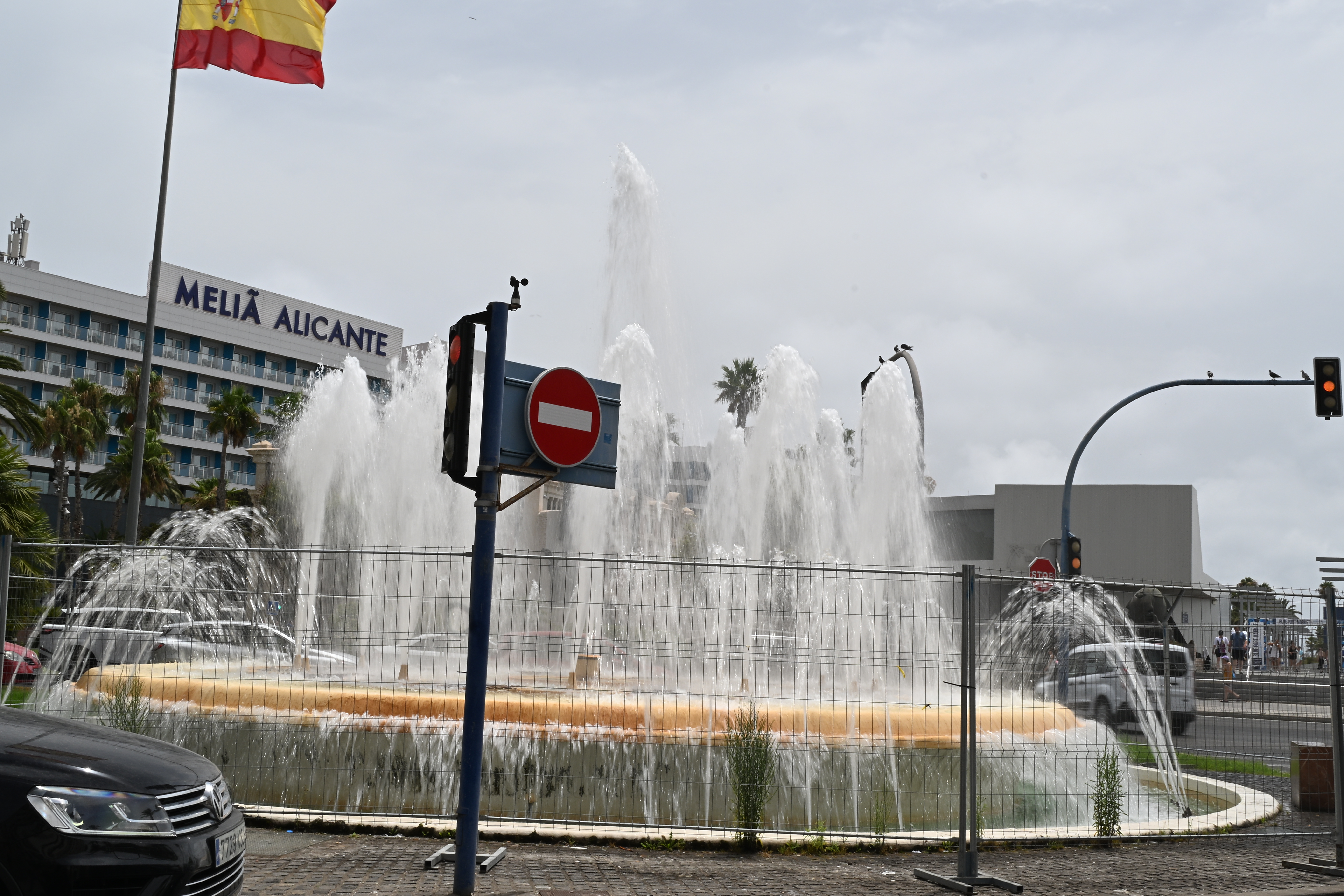 Wasserfontänen der Fuente de la Puerta del Mar hinter einer Absperrung, mit Verkehrszeichen, spanischer Flagge und Hotel in Alicante