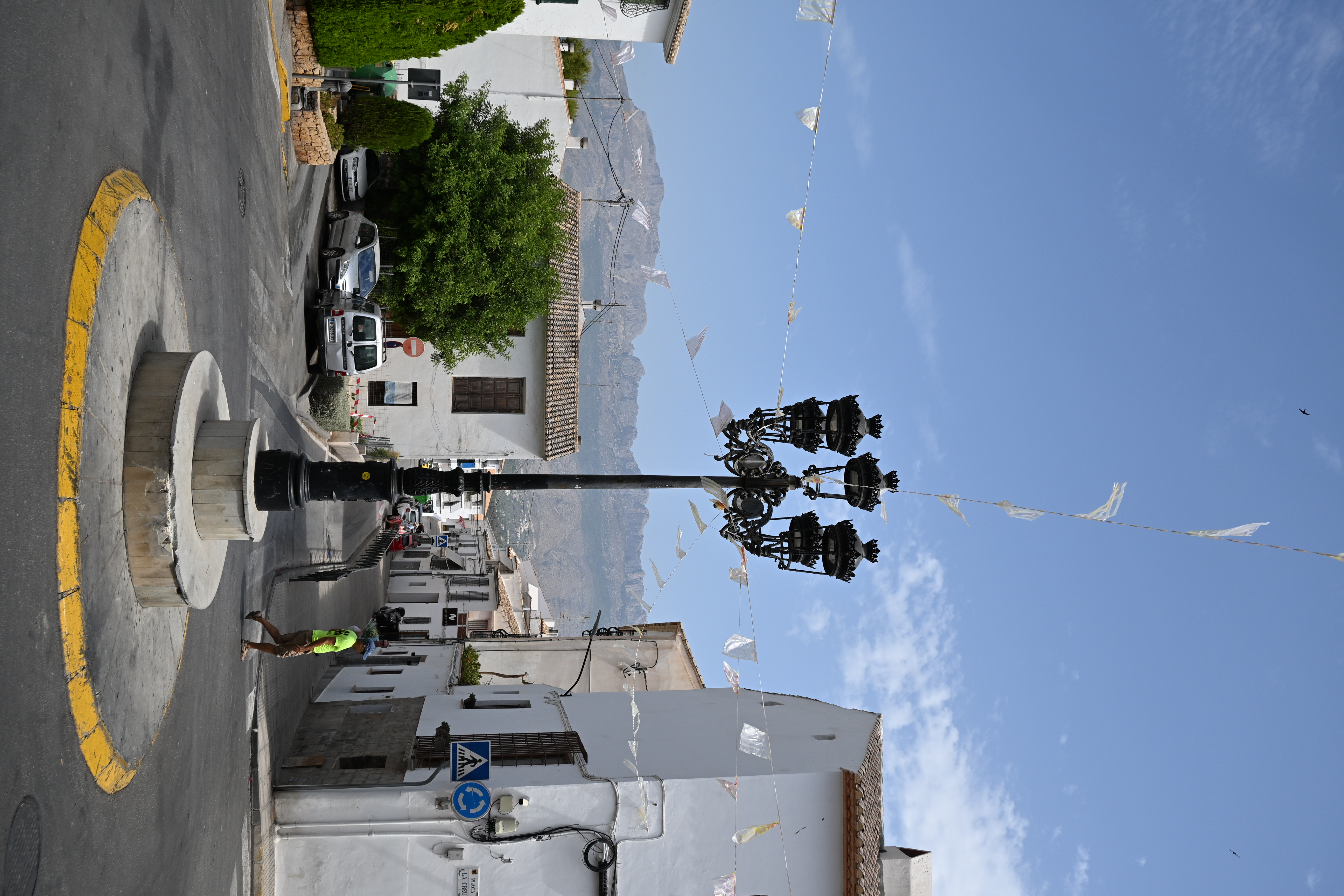 Verzierte schwarze Straßenlaterne an einer Kreuzung mit weißen Häusern, Wimpelketten und felsiger Bergkulisse in Altea.