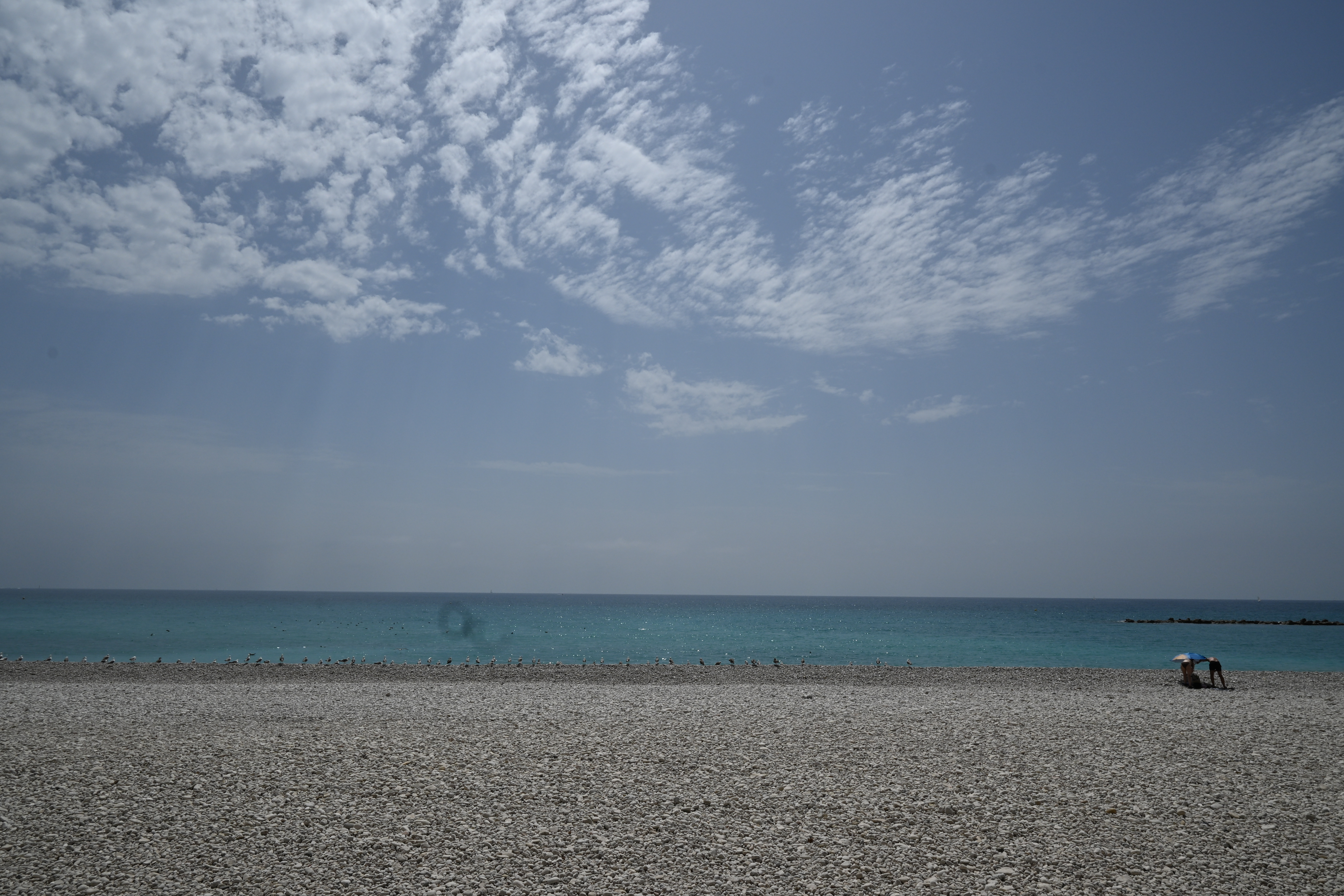 Kiesstrand der Playa de l’Espigó in Altea mit türkisfarbenem Meer, Vögeln am Ufer, zwei Personen und weitläufigem Wolkenhimmel.